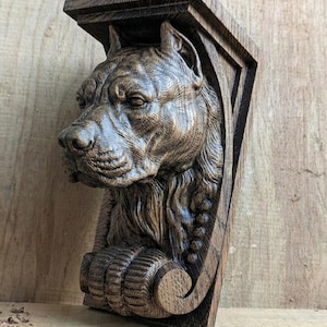 Puede incluir: Un soporte de madera tallada que representa la cabeza de un perro. La cara del perro está intrincadamente tallada con textura de pelaje visible y una expresión seria. El soporte es de color marrón oscuro y tiene un diseño arquitectónico decorativo.