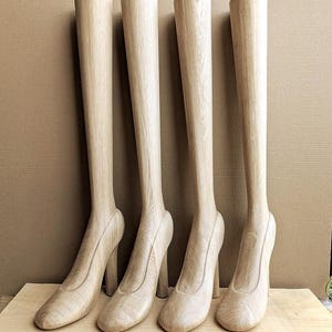 May include: Four light-colored wooden leg forms with attached high-heeled shoe shapes. Each form features a smooth, vertical grain pattern and a rounded toe design. These display pieces are arranged in a row, showcasing a natural wood finish.