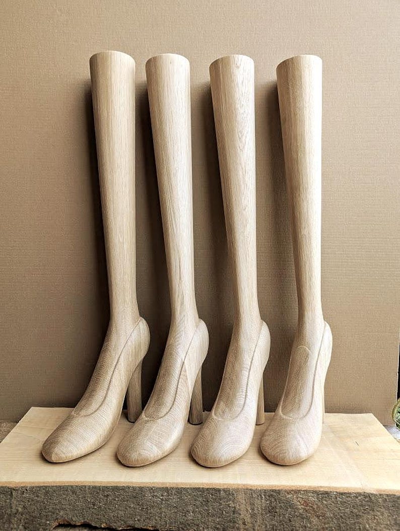 May include: Four light-colored wooden leg forms with attached high-heeled shoe shapes. Each form features a smooth, vertical grain pattern and a rounded toe design. These display pieces are arranged in a row, showcasing a natural wood finish.