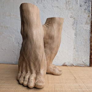 Op de afbeelding: Een paar lichtbruine houten sculpturen in de vorm van menselijke voeten en onderenkels. De houtsnijwerken vertonen zichtbare houtnerven, gedetailleerde teenstructuren en een gladde, natuurlijke afwerking, rechtopstaand als decoratieve objecten.
