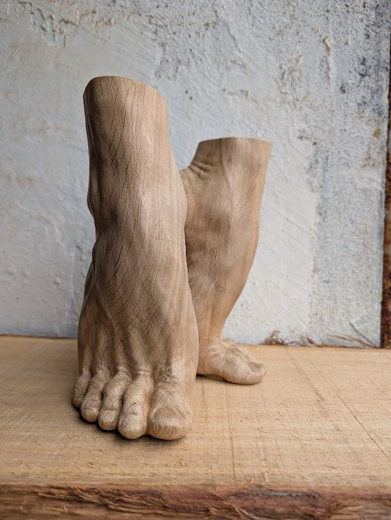 Op de afbeelding: Een paar lichtbruine houten sculpturen in de vorm van menselijke voeten en onderenkels. De houtsnijwerken vertonen zichtbare houtnerven, gedetailleerde teenstructuren en een gladde, natuurlijke afwerking, rechtopstaand als decoratieve objecten.