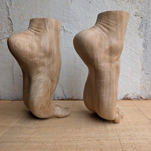 Op de afbeelding: Twee lichtbruine houten sculpturen uitgesneden in de vorm van menselijke voeten en enkels in een spitse positie. De houtnerf is zichtbaar over het oppervlak van beide objecten, wat de natuurlijke textuur en lichtbruine kleur van het materiaal toont.