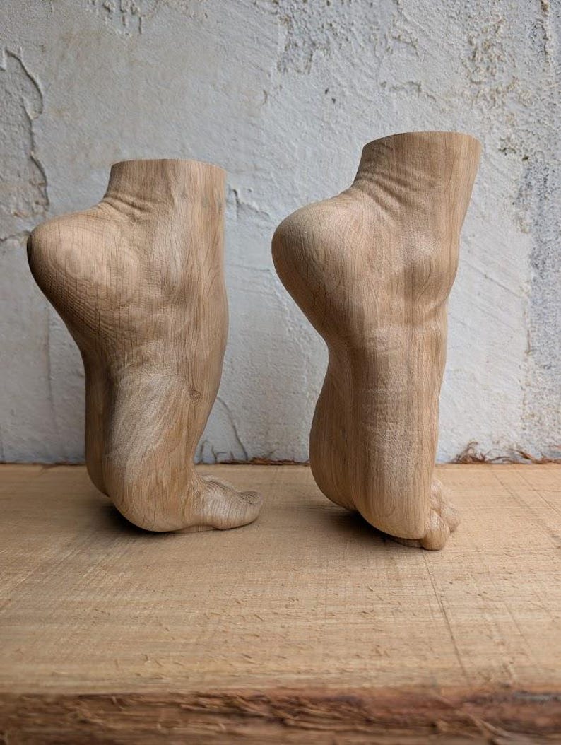 Op de afbeelding: Twee lichtbruine houten sculpturen uitgesneden in de vorm van menselijke voeten en enkels in een spitse positie. De houtnerf is zichtbaar over het oppervlak van beide objecten, wat de natuurlijke textuur en lichtbruine kleur van het materiaal toont.
