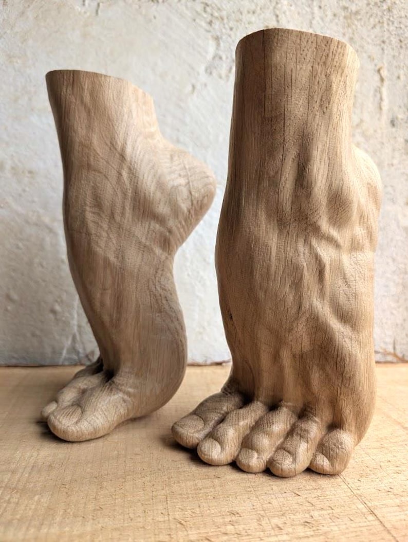 May include: Two light brown wooden sculptures carved into the shape of human feet and ankles. The carvings feature detailed textures representing skin, toes, and heel structures, with one foot shown from a side profile and the other from the front.