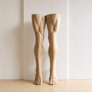 Pode incluir: Um par de pernas e pés de madeira em tom claro em posição vertical. A escultura apresenta entalhes anatômicos detalhados dos quadríceps, joelhos, panturrilhas e dedos, exibindo o veio natural e a textura da madeira em toda a sua forma.