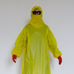 Könnte beinhalten: Eine Person trägt einen leuchtend gelben Ganzkörperanzug mit Kapuze und schwarzer Sonnenbrille. Der Anzug besteht aus einem glänzenden, wasserdichten Material. Die Person trägt schwarze und goldene Schuhe.