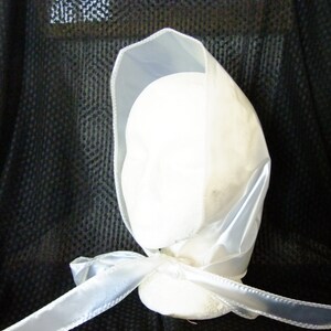 May include: A white satin bonnet with a white satin ribbon tied at the back. The bonnet is on a black background.