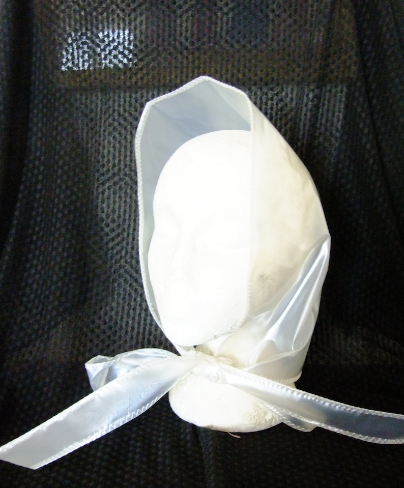 May include: A white satin bonnet with a white satin ribbon tied at the back. The bonnet is on a black background.