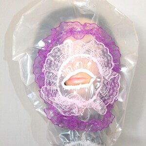 May include: A transparent plastic shower cap featuring a purple lace trim and a pink lace inner layer. The cap has a small mouth opening, revealing lips. The cap is worn over a person's head.
