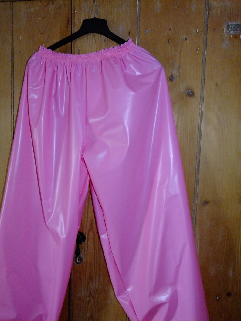 May include: Pink plastic pants hanging on a hanger. The pants have a gathered elastic waistband and are made of a shiny, smooth material.