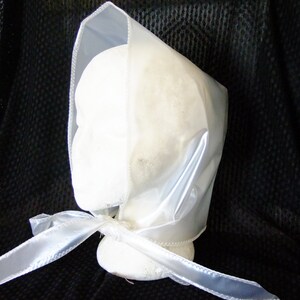 May include: A white satin bonnet with a sheer plastic overlay and a matching satin ribbon tie. The bonnet is displayed on a white mannequin head.