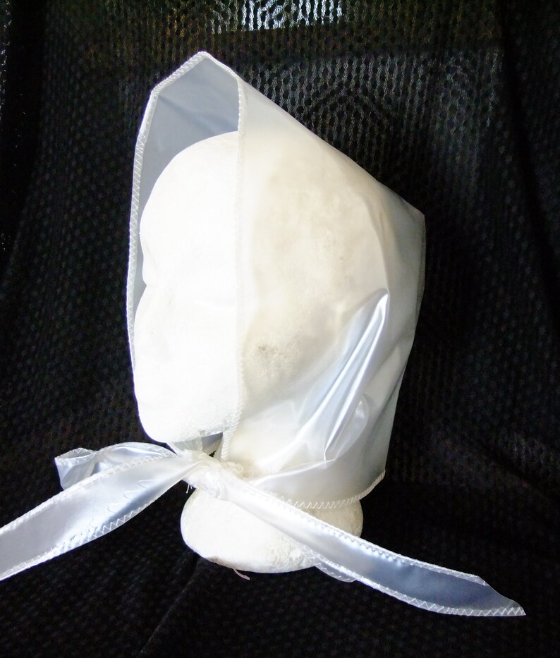 May include: A white satin bonnet with a sheer plastic overlay and a matching satin ribbon tie. The bonnet is displayed on a white mannequin head.
