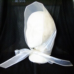 May include: A white satin bonnet with a bow tied at the back. The bonnet is made of a sheer fabric and has a simple design.