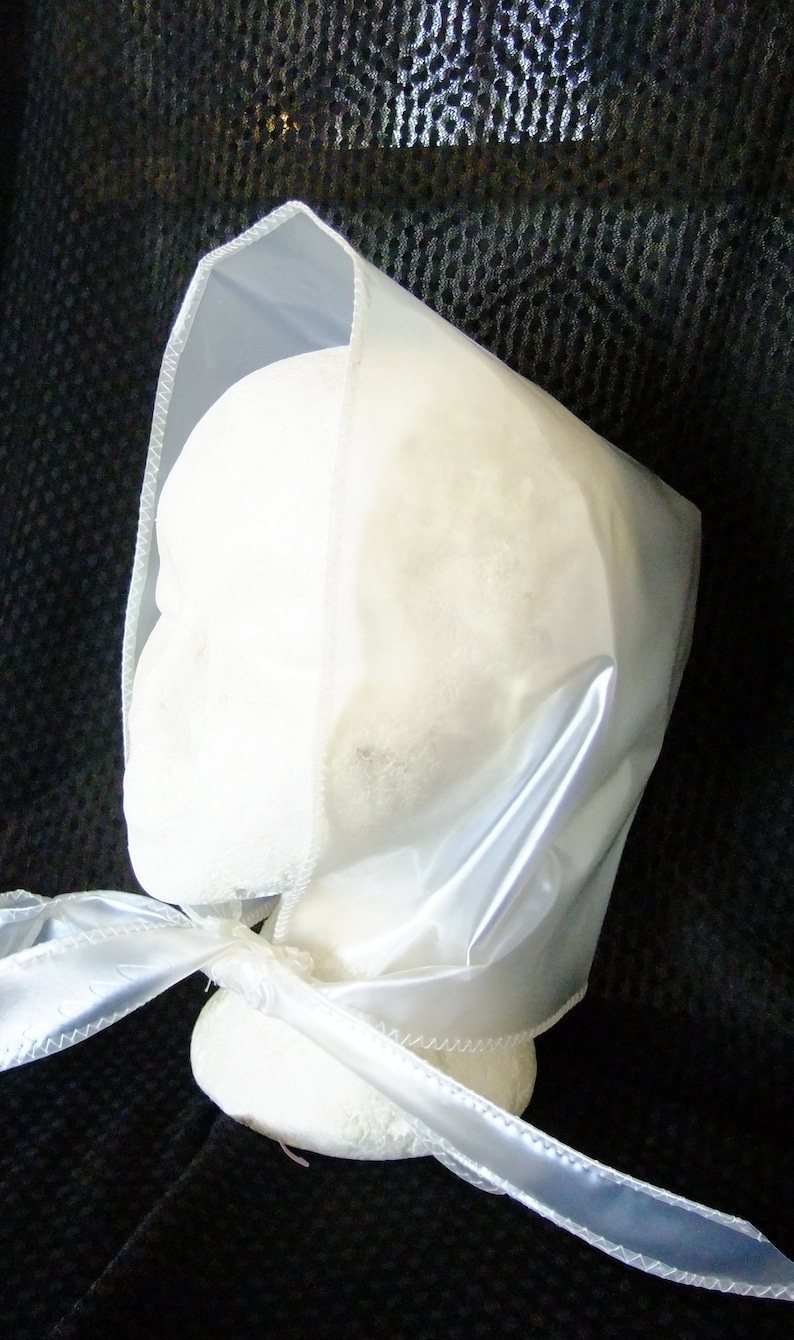 May include: A white satin bonnet with a gathered top and a long, flowing ribbon. The bonnet is made of a soft, silky fabric and has a delicate, feminine look.