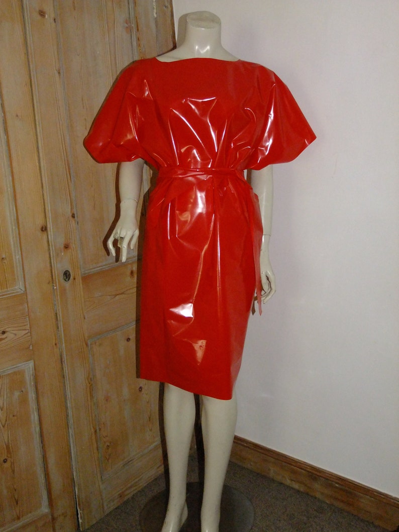 K&ouml;nnte beinhalten: Ein rotes, gl&auml;nzendes, &auml;rmelloses Kleid mit einem kurzen, figurbetonten Rock und einem breiten, roten G&uuml;rtel. Das Kleid ist aus einem gl&auml;nzenden Material gefertigt und hat ein schlichtes, elegantes Design.