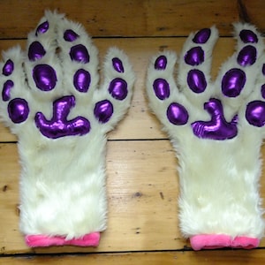 May include: A pair of white furry paw gloves with purple paw pads. The gloves have pink cuffs.