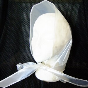 May include: A white satin bonnet with a white satin ribbon tied at the back. The bonnet is made of a sheer fabric and has a simple design.