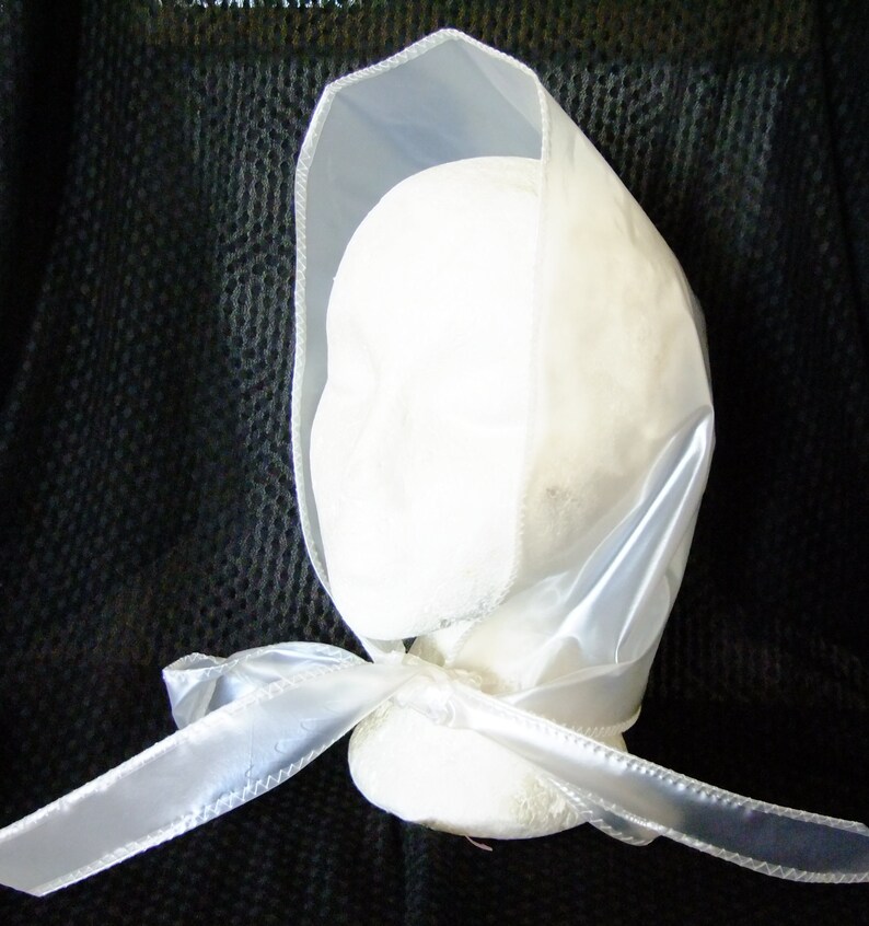May include: A white satin bonnet with a white satin ribbon tied at the back. The bonnet is made of a sheer fabric and has a simple design.