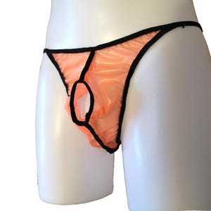 Könnte beinhalten: Pfirsichfarbener String-Tanga mit schwarzem Besatz und Schlüsselloch-Design. Die Unterwäsche wird auf einer weißen Schaufensterpuppe präsentiert, die die Form und Details des Kleidungsstücks zeigt. Der Stoff scheint halbtransparent zu sein.