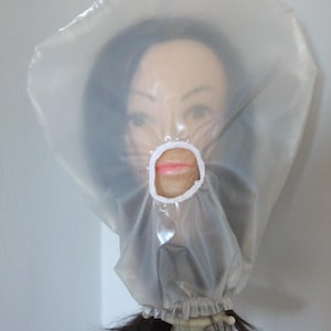 May include: A transparent plastic hood with a white trim around the mouth area, covering a mannequin head. The mannequin has dark hair and is mounted on a black stand. The hood is translucent, allowing the face to be partially visible.