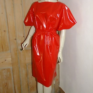 K&ouml;nnte beinhalten: Ein rotes, gl&auml;nzendes, &auml;rmelloses Kleid mit einem kurzen, weiten Ausschnitt und einem G&uuml;rtel. Das Kleid ist aus einem glatten, gl&auml;nzenden Material gefertigt.