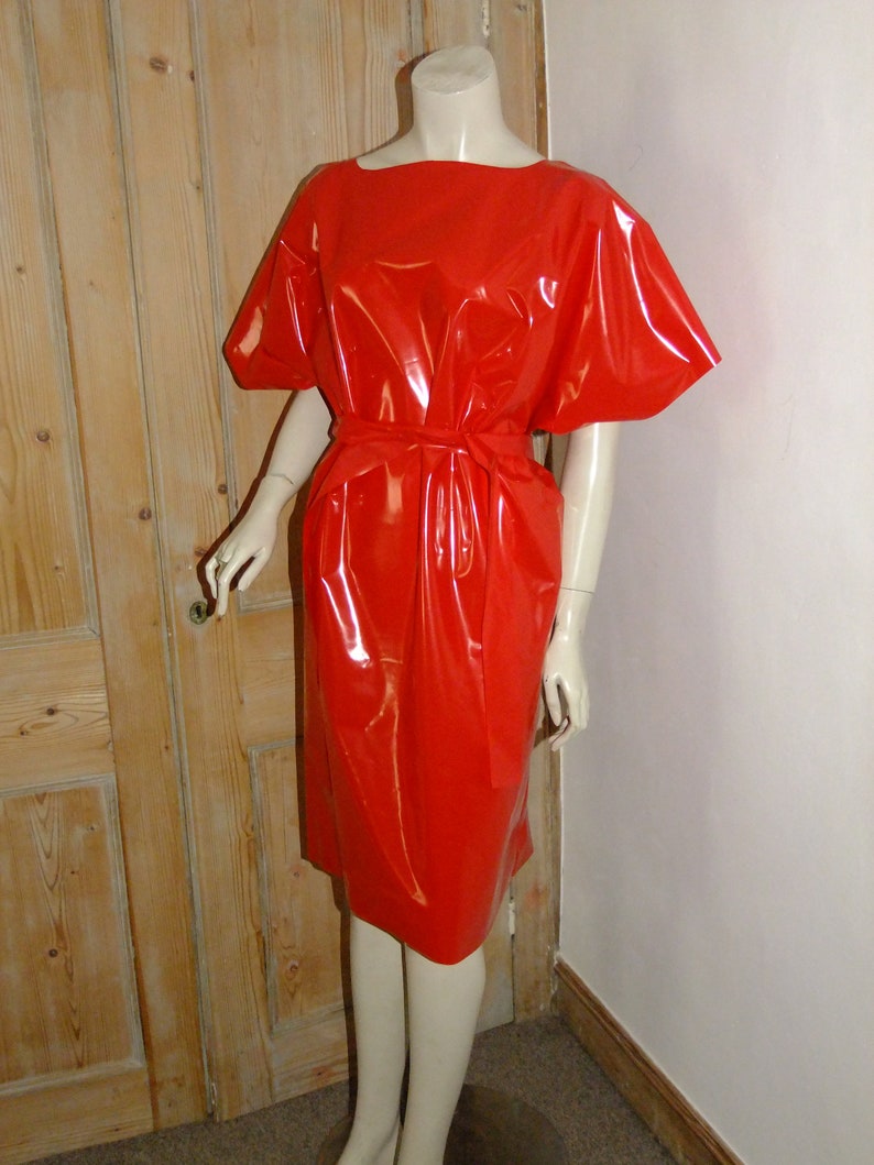 K&ouml;nnte beinhalten: Ein rotes, gl&auml;nzendes, &auml;rmelloses Kleid mit einem kurzen, weiten Ausschnitt und einem G&uuml;rtel. Das Kleid ist aus einem glatten, gl&auml;nzenden Material gefertigt.
