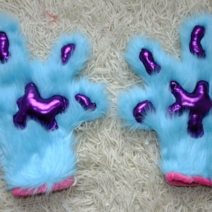 Puede incluir: Un par de títeres de mano de peluche azul con almohadillas de patas moradas. Los títeres tienen puños rosas.