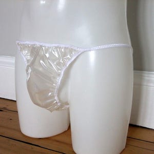 May include: A white, transparent thong-style underwear with white trim. The underwear is made of a shiny, plastic-like material. The garment is displayed on a white mannequin.