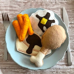 May include: A light blue plate with a felt meal including a drumstick, carrots, a pancake with butter, and a slice of bread with gravy. A fork and knife are on either side of the plate.