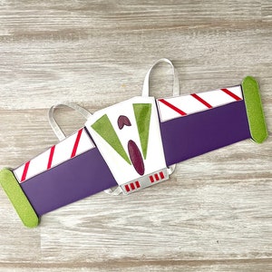 May include: A white, green, and purple felt costume wing with red stripes and silver accents. The wing is shaped like a spaceship and has a green and white face on the front.