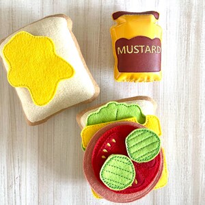 May include: Felt toy food set with two slices of bread, one with yellow mustard, one with lettuce, tomato, and pickles, and a jar of mustard labeled "MUSTARD".