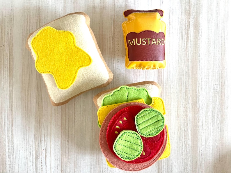 May include: Felt toy food set with two slices of bread, one with yellow mustard, one with lettuce, tomato, and pickles, and a jar of mustard labeled "MUSTARD".