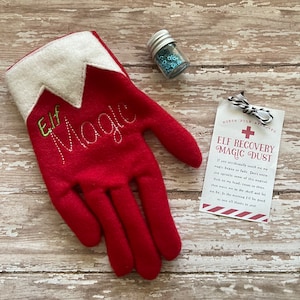 May include: Red felt elf glove with white trim and the words "Elf Magic" embroidered on it. A small jar of blue glitter and a note that reads "North Pole Approved + Elf Recovery Magic Dust" is next to the glove.