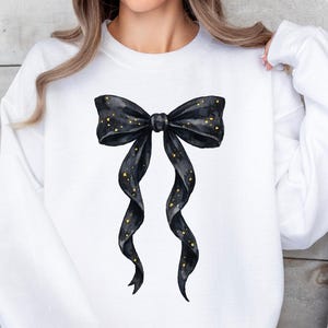 May include: White sweatshirt featuring a large black bow with gold glitter accents. The bow is centered on the front of the long-sleeved top. The bow's ribbons cascade downwards, adding a touch of elegance to the casual wear.