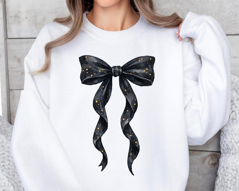 May include: White sweatshirt featuring a large black bow with gold glitter accents. The bow is centered on the front of the long-sleeved top. The bow's ribbons cascade downwards, adding a touch of elegance to the casual wear.