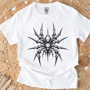 May include: White t-shirt featuring a detailed black and gray spider graphic. The spider design has sharp, spiky legs and a central body, creating a striking contrast against the white fabric. The shirt is a classic crew neck style.