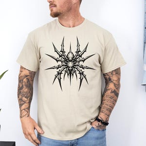 May include: A beige t-shirt featuring a detailed black and white spider graphic on the front. The spider design has sharp, spiky legs radiating from a central body. The shirt is a short-sleeved crew neck.