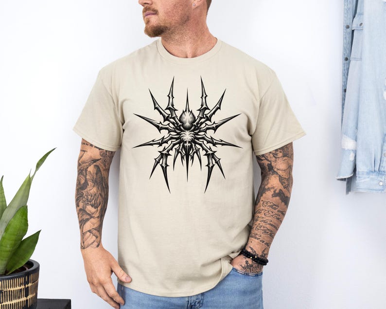 May include: A beige t-shirt featuring a detailed black and white spider graphic on the front. The spider design has sharp, spiky legs radiating from a central body. The shirt is a short-sleeved crew neck.
