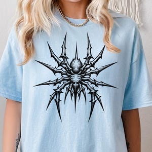 May include: A light blue t-shirt featuring a detailed black and white spider graphic. The spider design has sharp, spiky legs radiating from a central body. The shirt is a casual, comfortable style.