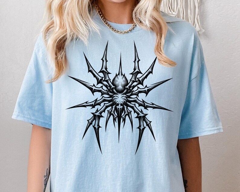 May include: A light blue t-shirt featuring a detailed black and white spider graphic. The spider design has sharp, spiky legs radiating from a central body. The shirt is a casual, comfortable style.