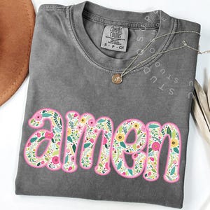 Op de afbeelding: Een grijs T-shirt met het woord "amen" in roze letters. De letters zijn gevuld met een bloemenpatroon in tinten roze, geel en groen.