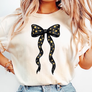 May include: Cream-colored t-shirt featuring a black bow design adorned with gold stars. The bow has long, flowing ribbons with star accents. The design is centered on the shirt, creating a visually appealing graphic.
