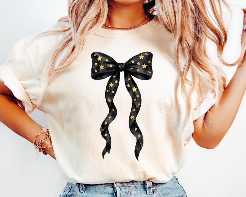 May include: Cream-colored t-shirt featuring a black bow design adorned with gold stars. The bow has long, flowing ribbons with star accents. The design is centered on the shirt, creating a visually appealing graphic.
