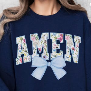 Op de afbeelding: Een marineblauw sweatshirt met het woord "AMEN" in witte letters omringd door een bloemenpatroon. Een lichtblauwe strik is gecentreerd onder het woord.