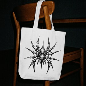 May include: White tote bag featuring a detailed black and gray spider design. The spider has sharp, spiky legs radiating from its body. The bag has a simple, rectangular shape with white handles, and is hanging on a wooden chair.