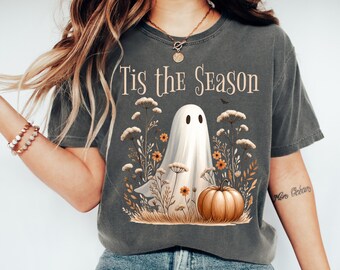 May include: Dark grey t-shirt featuring the text "Tis the Season" above a ghost illustration. The ghost is surrounded by flowers and a pumpkin. A bat is flying above the ghost. The shirt is a casual style.