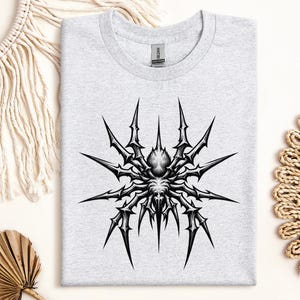 May include: A light gray t-shirt featuring a detailed black and white spider graphic. The spider design has sharp, spiky legs radiating from a central body. The shirt is folded neatly on a neutral surface.