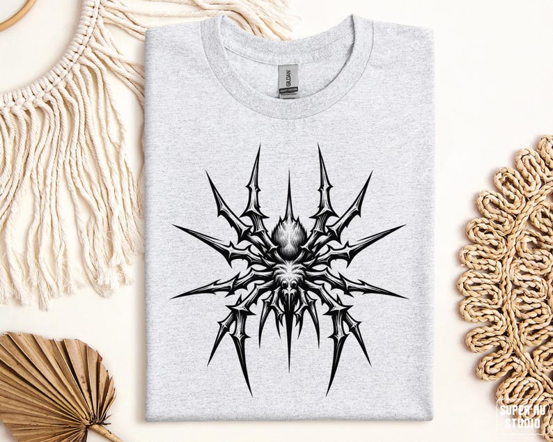 May include: A light gray t-shirt featuring a detailed black and white spider graphic. The spider design has sharp, spiky legs radiating from a central body. The shirt is folded neatly on a neutral surface.