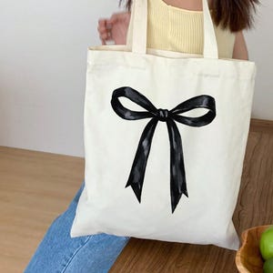 May include: A cream-colored tote bag featuring a large, black bow design. The bow is centered on the bag and has a watercolor-like appearance. The bag has two handles and appears to be made of canvas or a similar fabric.