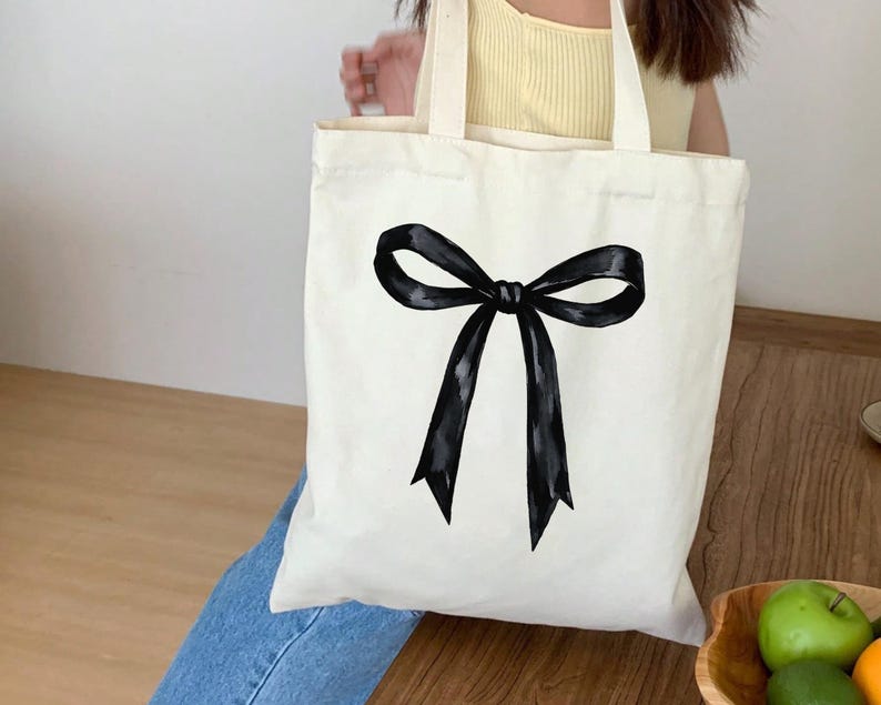 May include: A cream-colored tote bag featuring a large, black bow design. The bow is centered on the bag and has a watercolor-like appearance. The bag has two handles and appears to be made of canvas or a similar fabric.